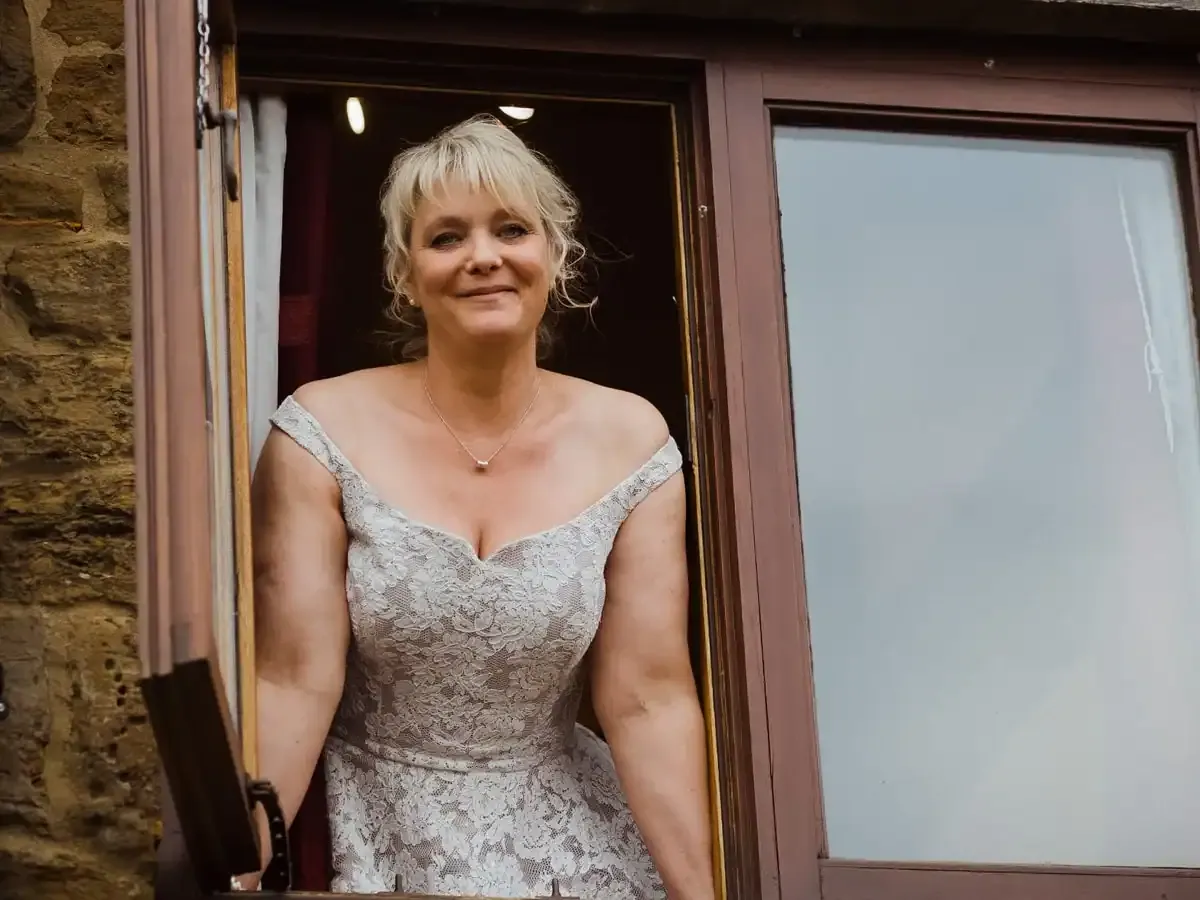 Blonde woman in a lace dress smiling from an open window.