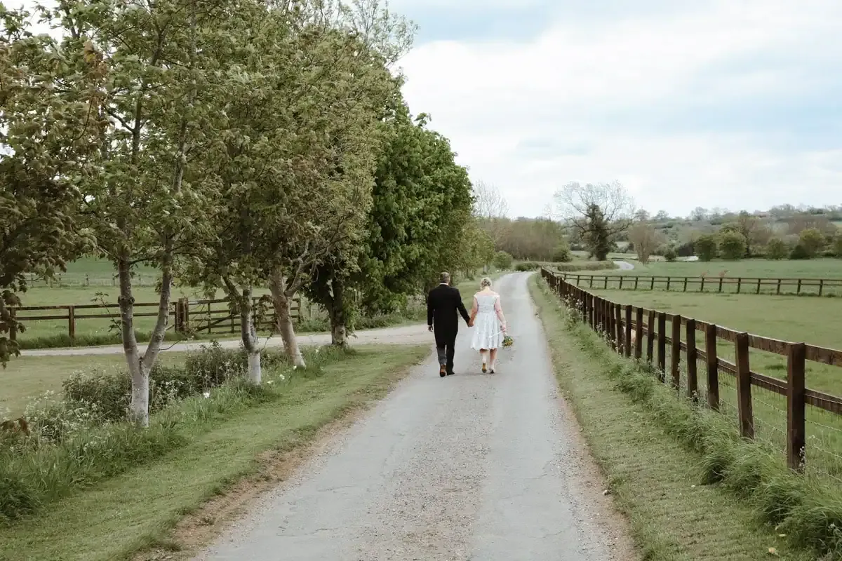 Couple holding hands walking down a country lane with trees and fields.