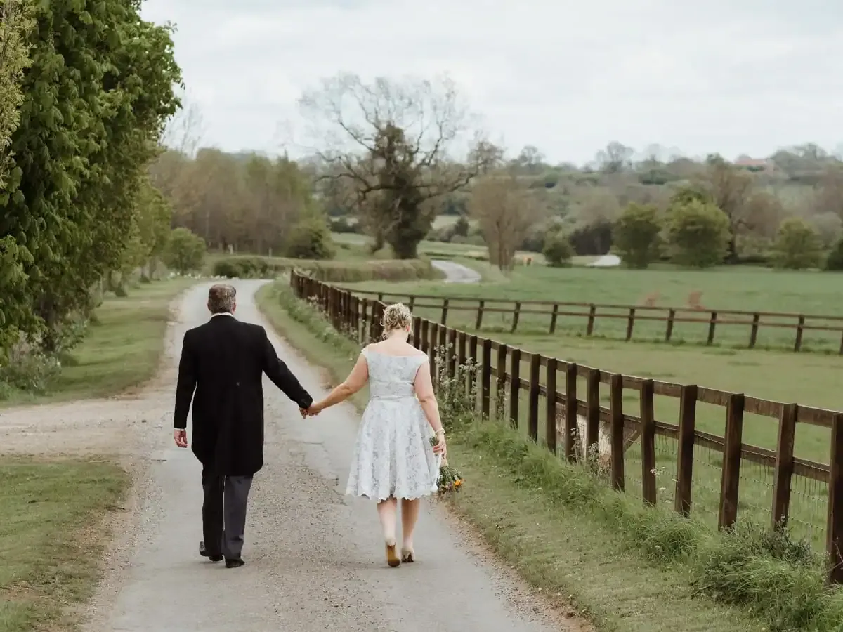Man in suit, woman in dress holding hands on a country path by a fence.