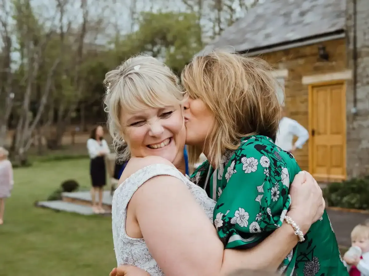 Smiling woman in white dress receives a kiss from woman in green floral.