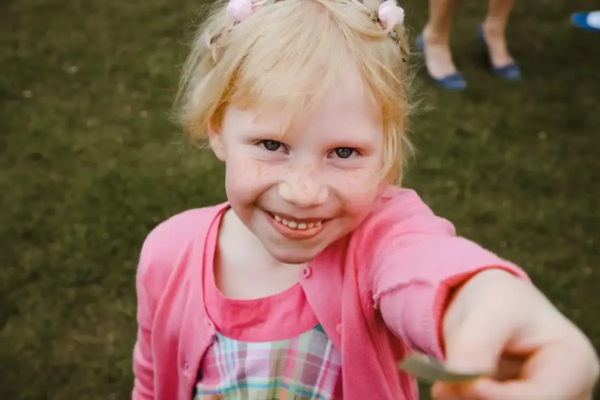 Smiling blonde girl with freckles offering a leaf, wearing pink and a floral headband.