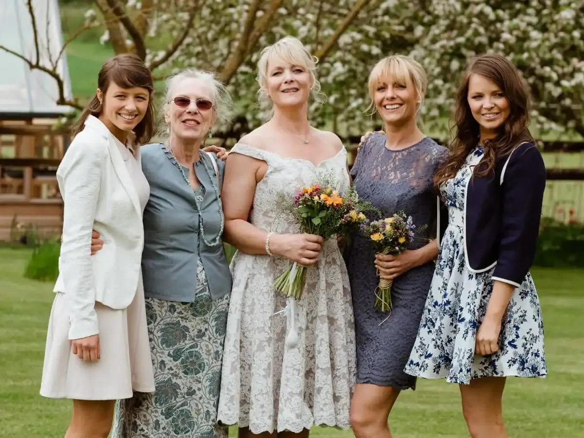 Five smiling women of varying ages outdoors, two holding flower bouquets.
