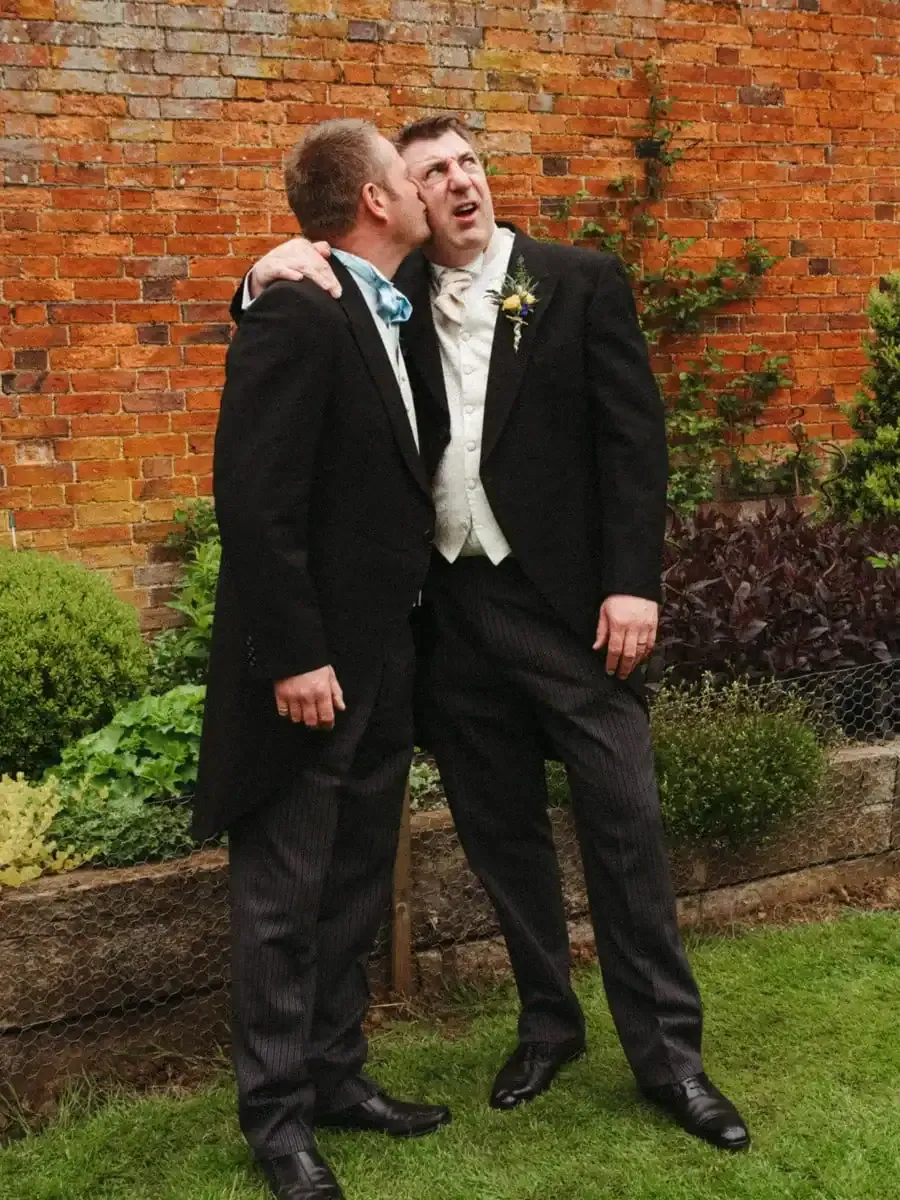 Man in morning suit kisses another man's cheek, who looks surprised.