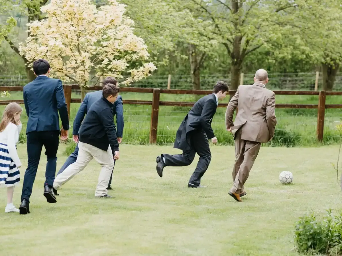 People in suits and casual clothes playing football on a grassy lawn.