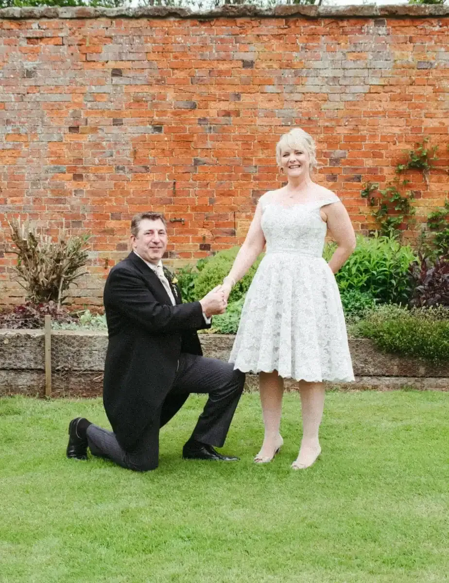 Man in suit kneels, holding hands with woman in white dress on grass.