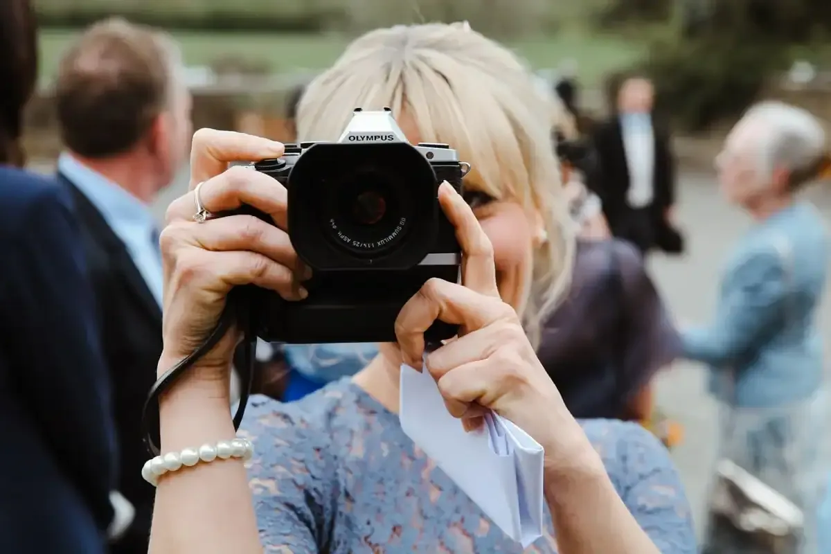 Blonde woman holding Olympus camera, pearl bracelet, looking at viewer.