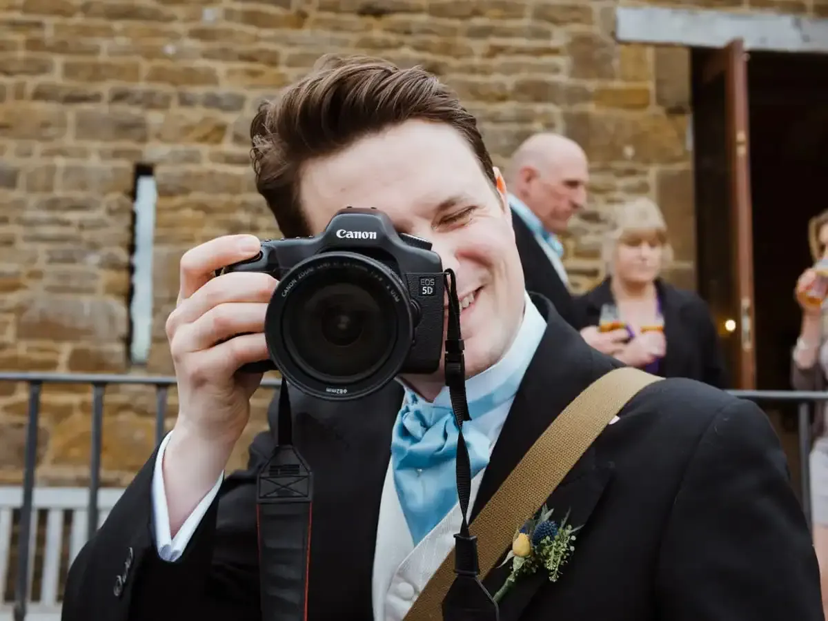 Smiling man in suit holding Canon EOS 5D camera to eye.