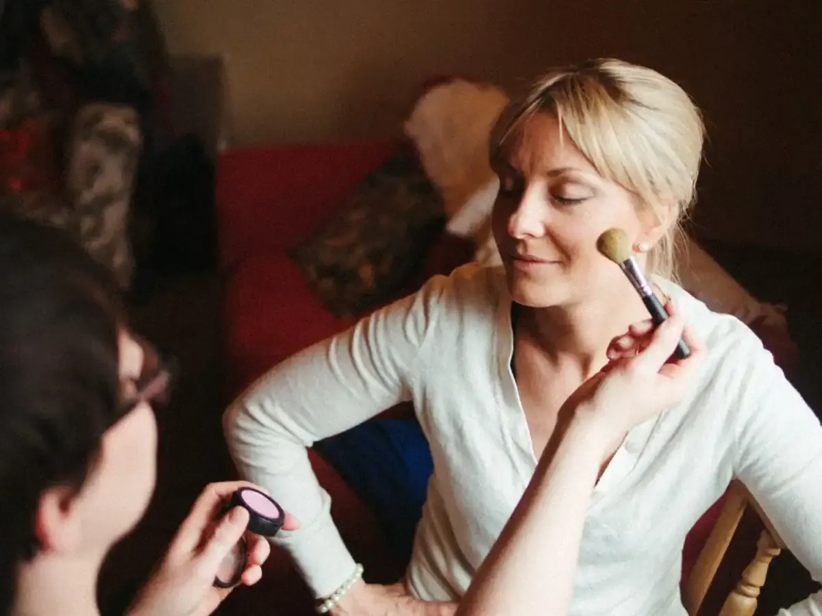 Hands apply makeup to blonde woman's cheek with brush and pink compact.