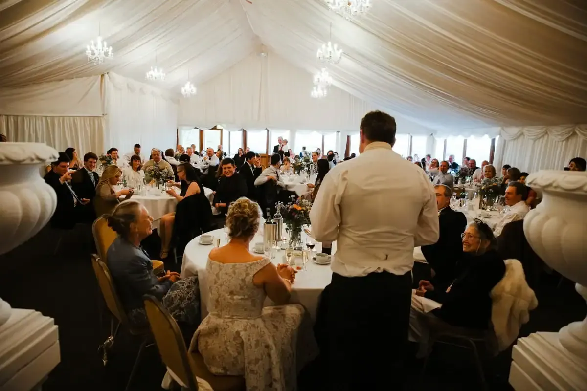 A man speaks to guests seated at tables in a white marquee.