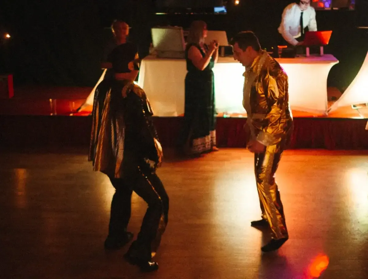 Two men dancing, one in a gold suit, the other in a shiny cape.
