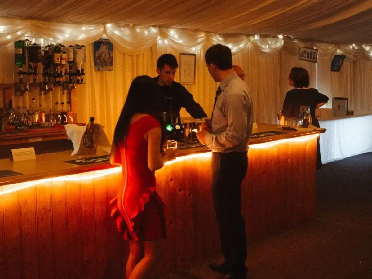 Bartender serves a woman in red and a man at a glowing wooden bar.