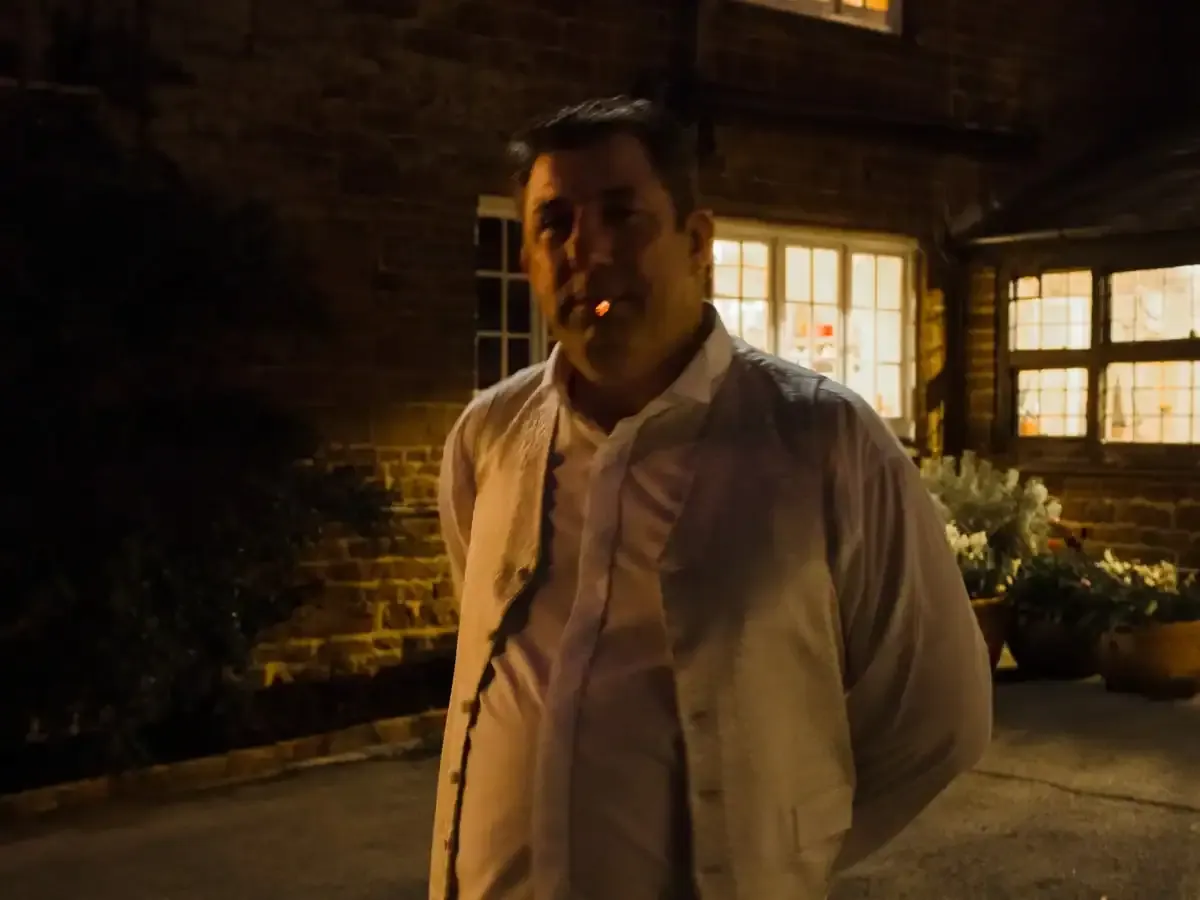 Man in waistcoat smoking outside a lit brick building at night.