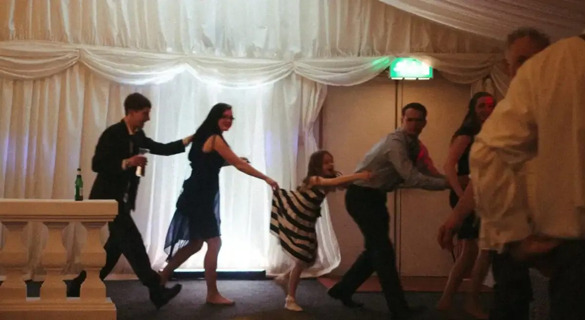 Guests of all ages dancing in a line, holding onto each other at a party.
