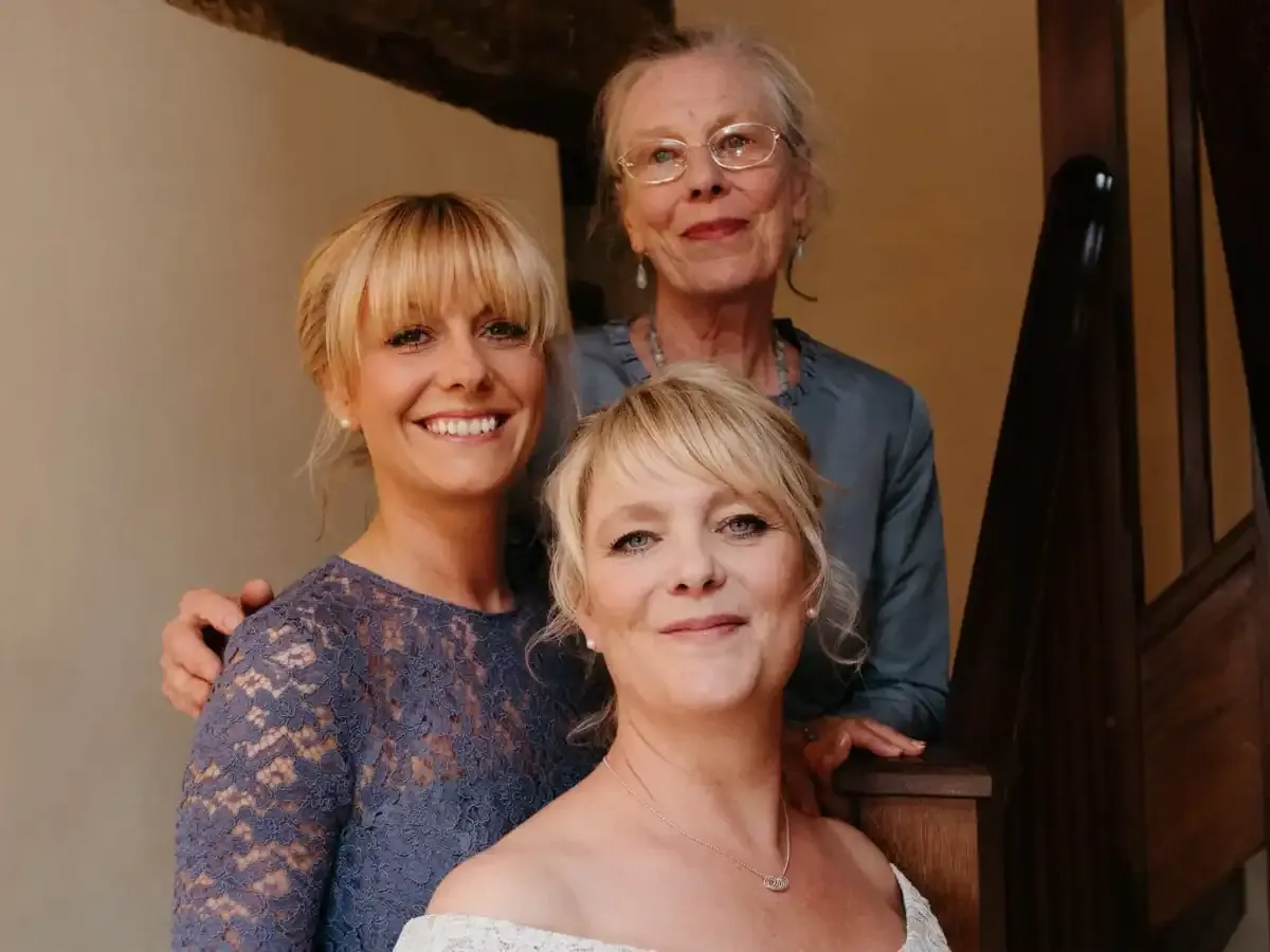 Three smiling women, different generations, by a wooden staircase.
