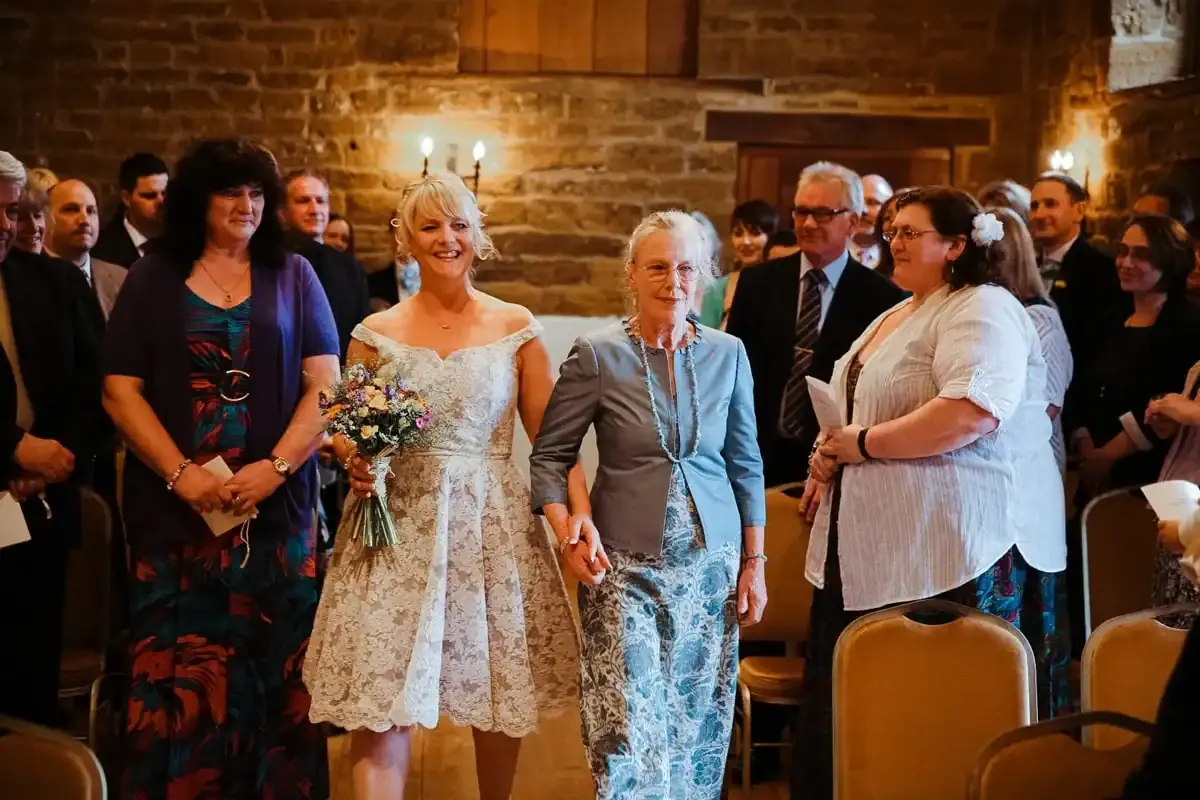 Smiling bride with bouquet walks down aisle holding older woman's hand.