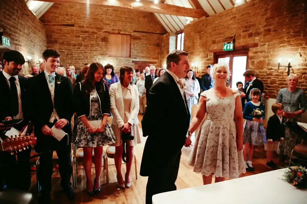 Smiling bride and groom holding hands with guests in a stone-walled barn.