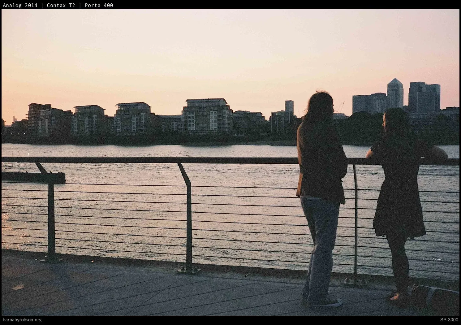 Two figures silhouetted at a riverside railing at dusk, shot on film.