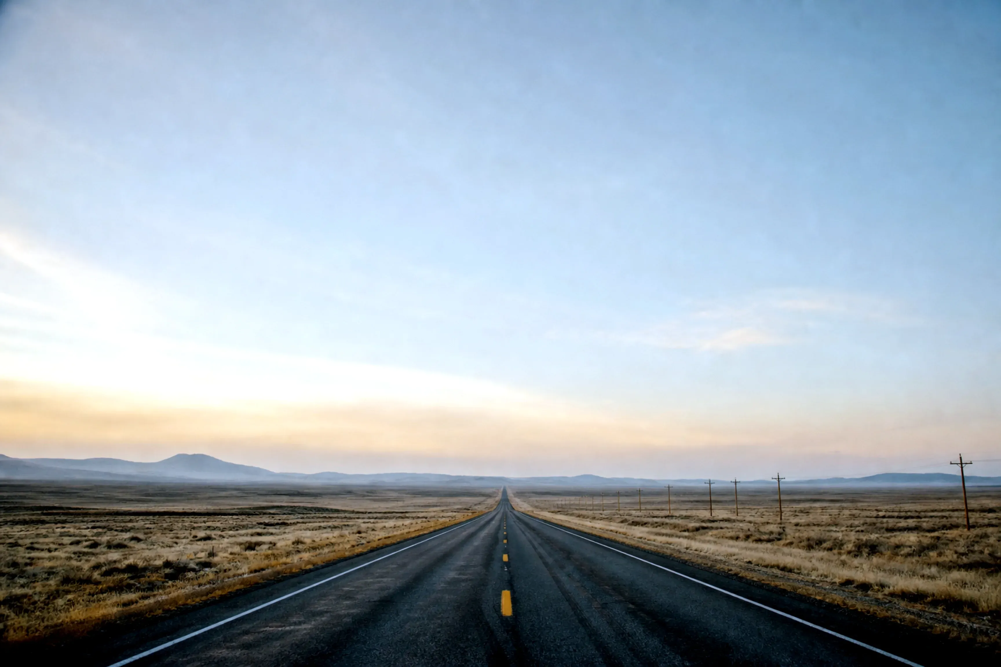An empty highway running straight to a distant horizon under a vast pale dawn sky, open plains stretching away on either side.