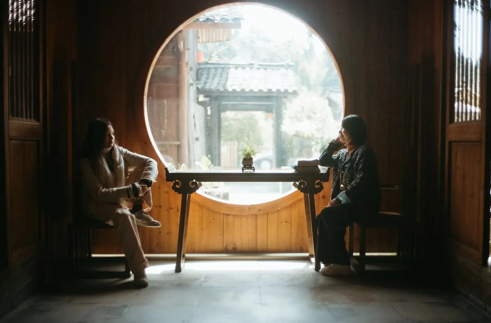Two people talking beside a round moon-gate window in a wooden teahouse