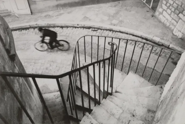 A cyclist blurring past below a spiral staircase railing, black and white.