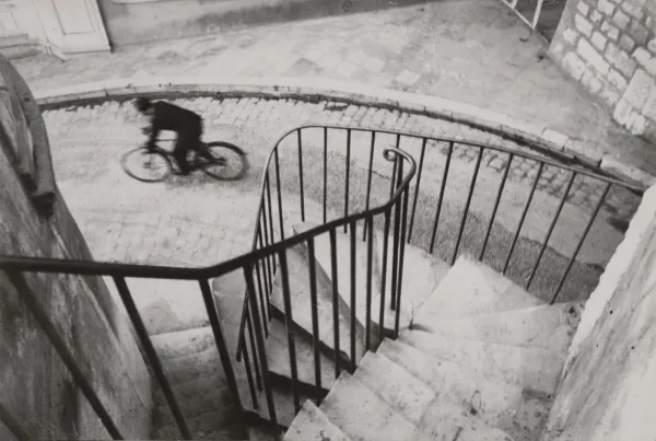 A cyclist blurring past below a spiral staircase railing, black and white.