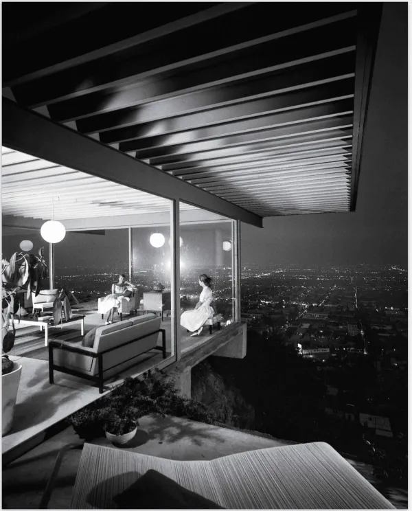 A glass-walled room cantilevered over a city at night, two women seated inside beneath a low ceiling, the lights of the grid spread out below.