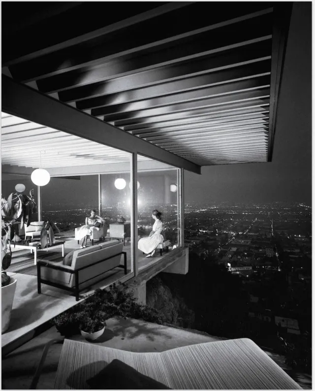 A glass-walled room cantilevered over a city at night, two women seated inside beneath a low ceiling, the lights of the grid spread out below.