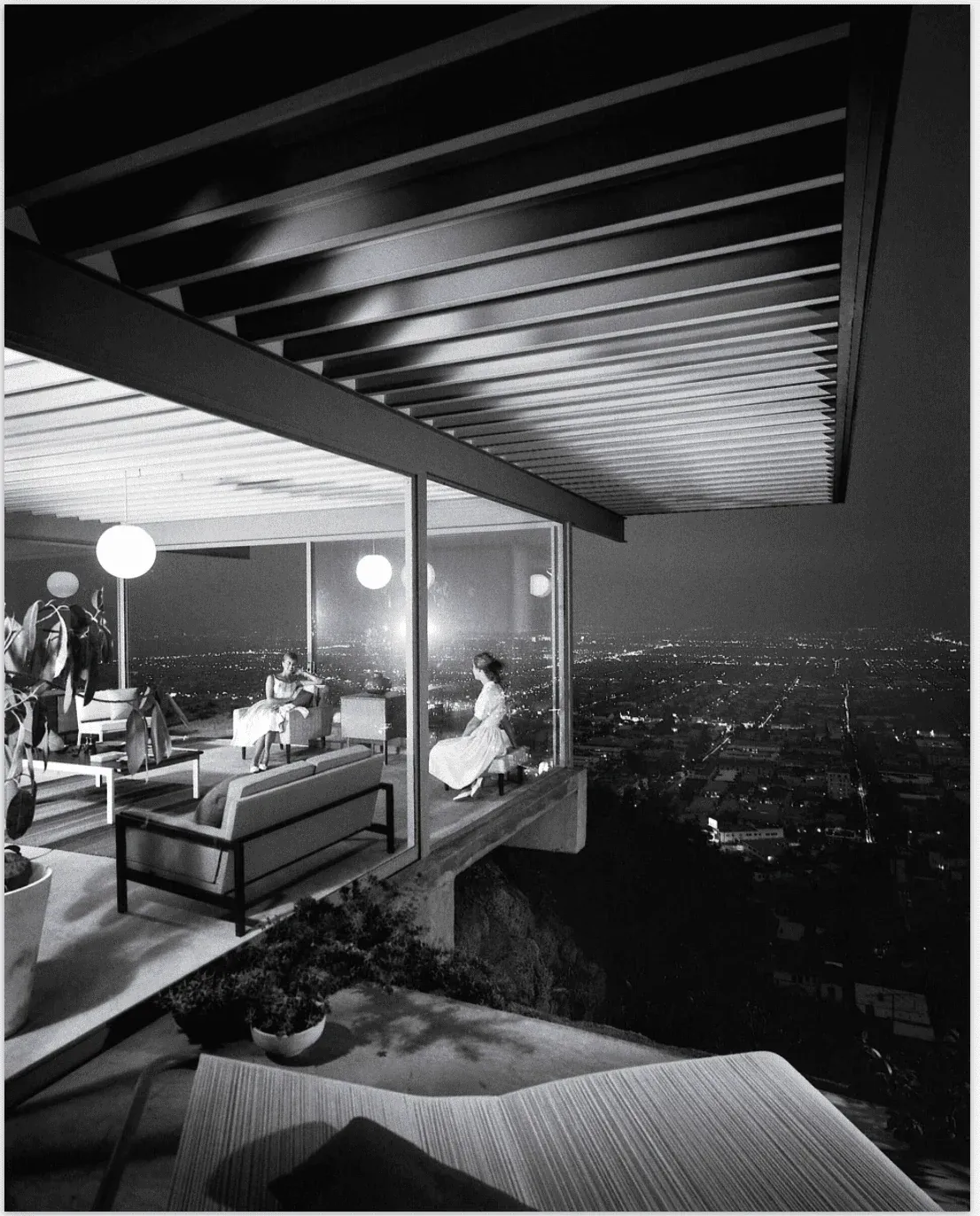 A glass-walled room cantilevered over a city at night, two women seated inside beneath a low ceiling, the lights of the grid spread out below.