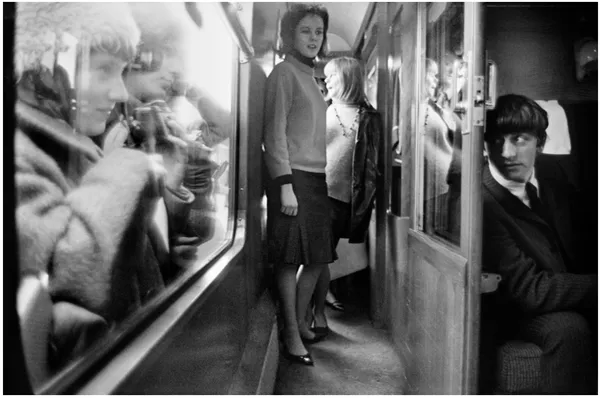 Black and white: Man seated, two women standing, reflection in train corridor.