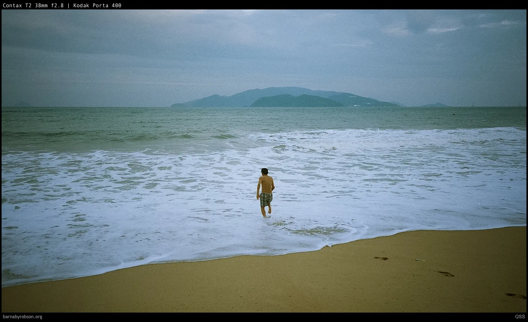 Man walking into the sea with waves, sandy beach, and distant hills.