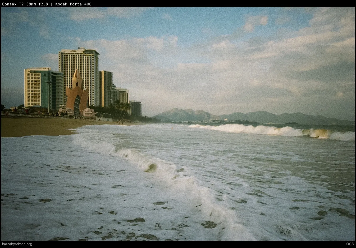 Waves breaking on a sandy beach with city buildings and mountains.