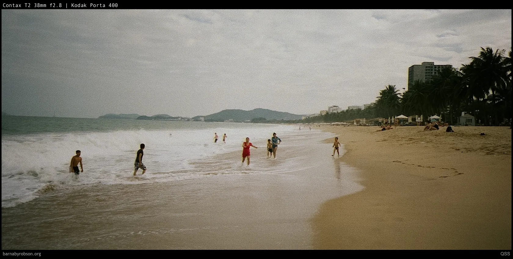 Holidaymakers enjoying a sandy beach with waves, palm trees, and distant buildings.