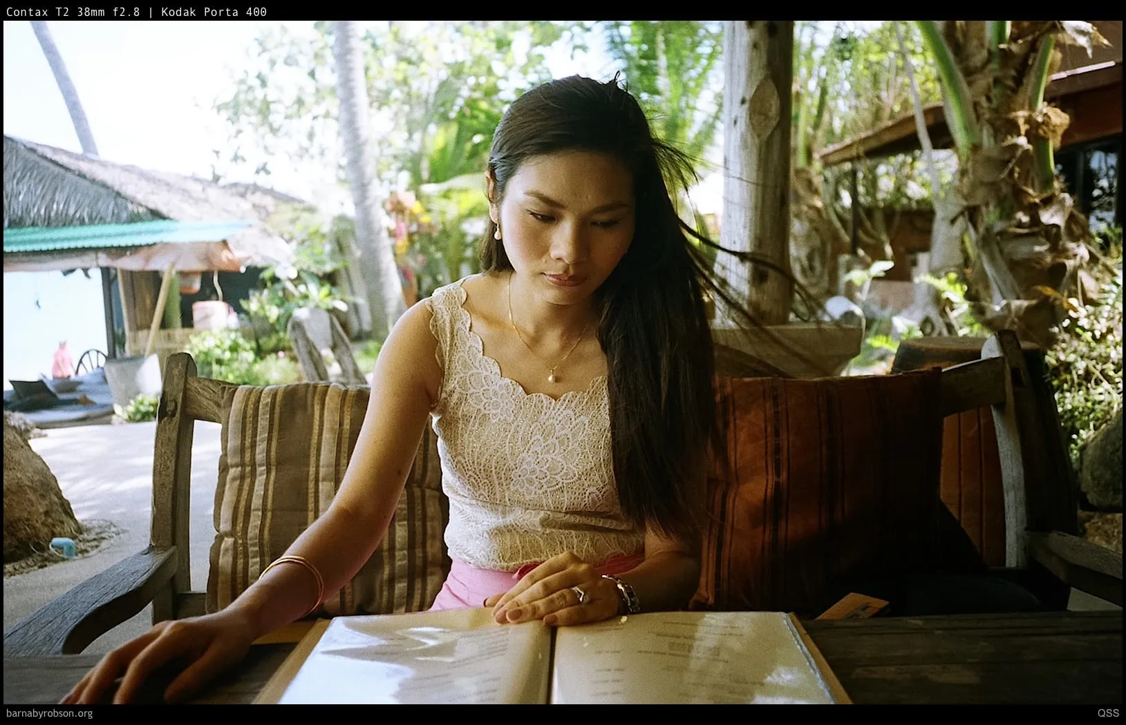 Woman in white lace top reading menu at tropical outdoor cafe.