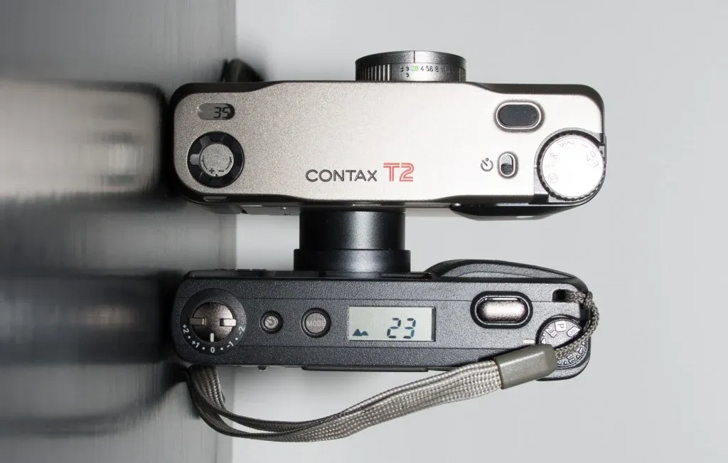 The Contax T2 and Ricoh GR1v seen from above, one stacked over the other — the T2's silver top plate and the GR1v's black one, frame counter reading 23.