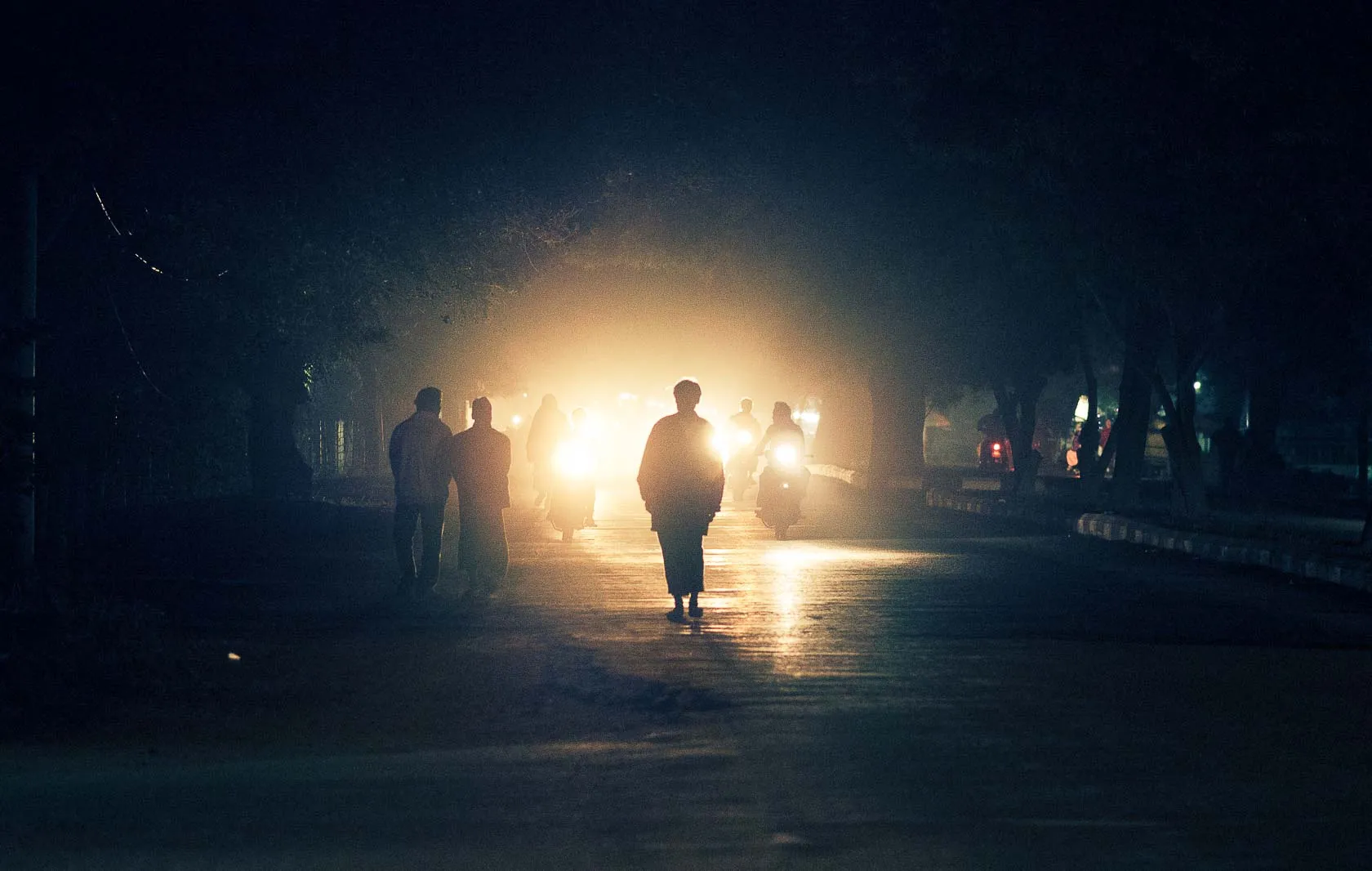 Silhouetted people and motorbikes on a dark road, illuminated by bright headlights.