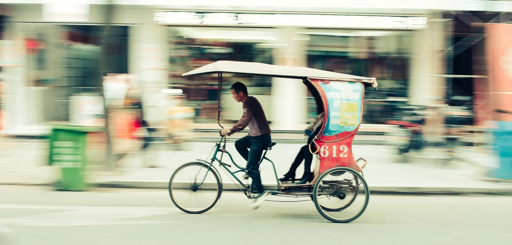 Man pedals red rickshaw with passenger, '612' visible, blurred street.