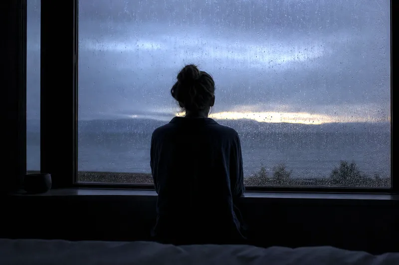 A figure at a rain-streaked window at dawn watching a thin break of golden light through grey cloud.