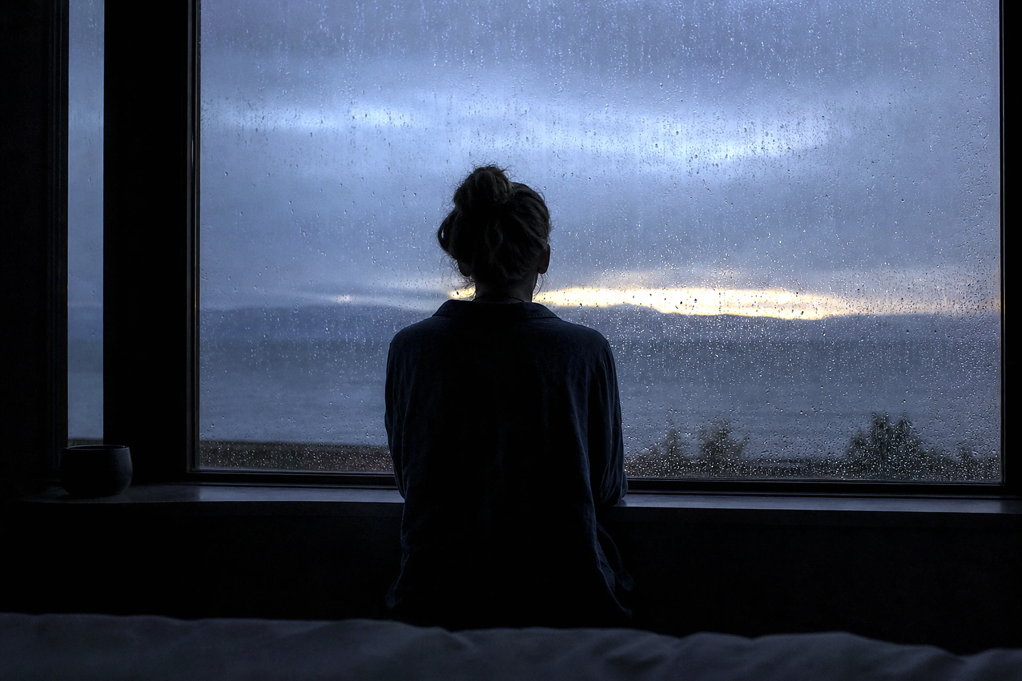 A figure at a rain-streaked window at dawn watching a thin break of golden light through grey cloud.