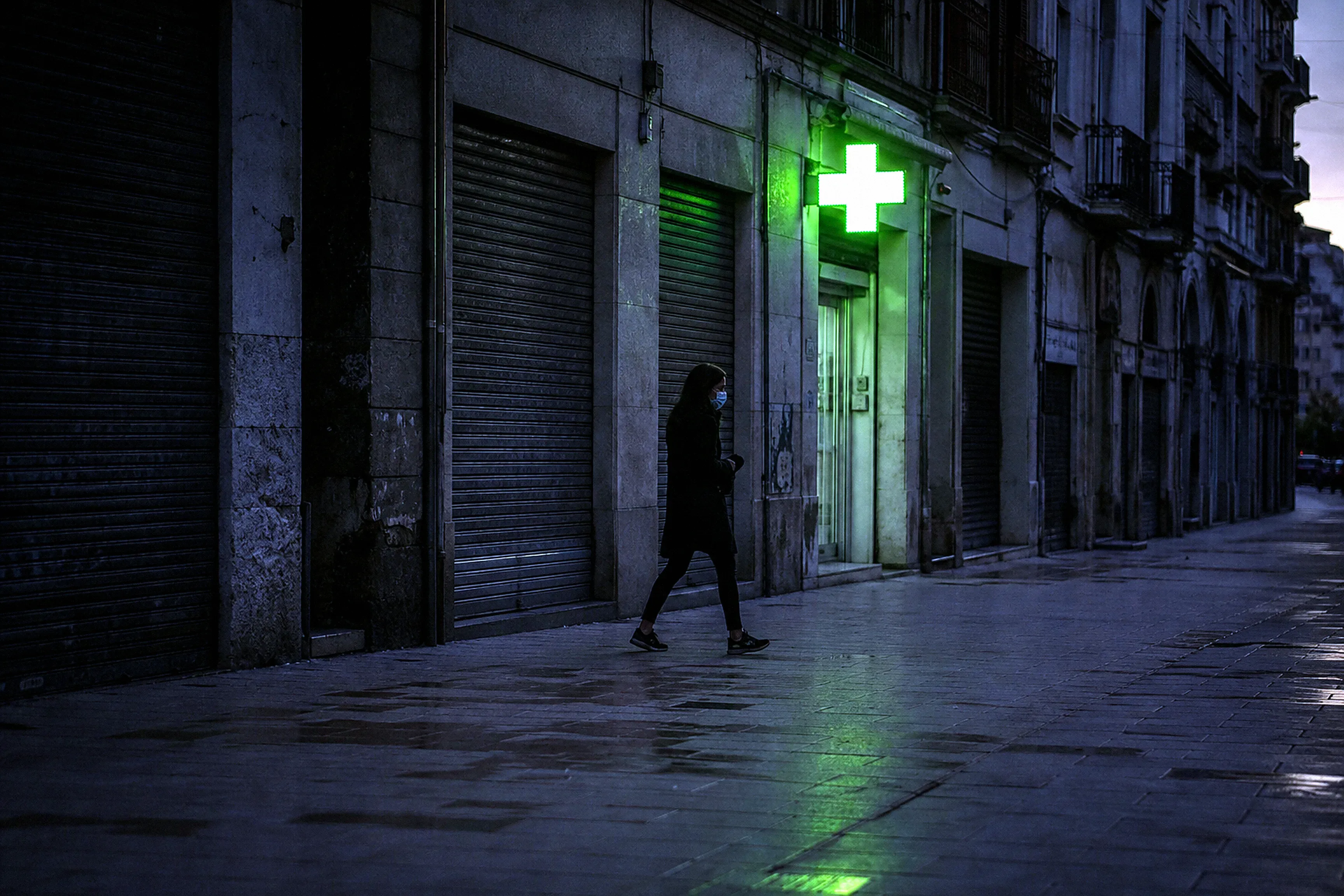 A lone masked figure passing the one glowing pharmacy cross on a shuttered street at dusk.