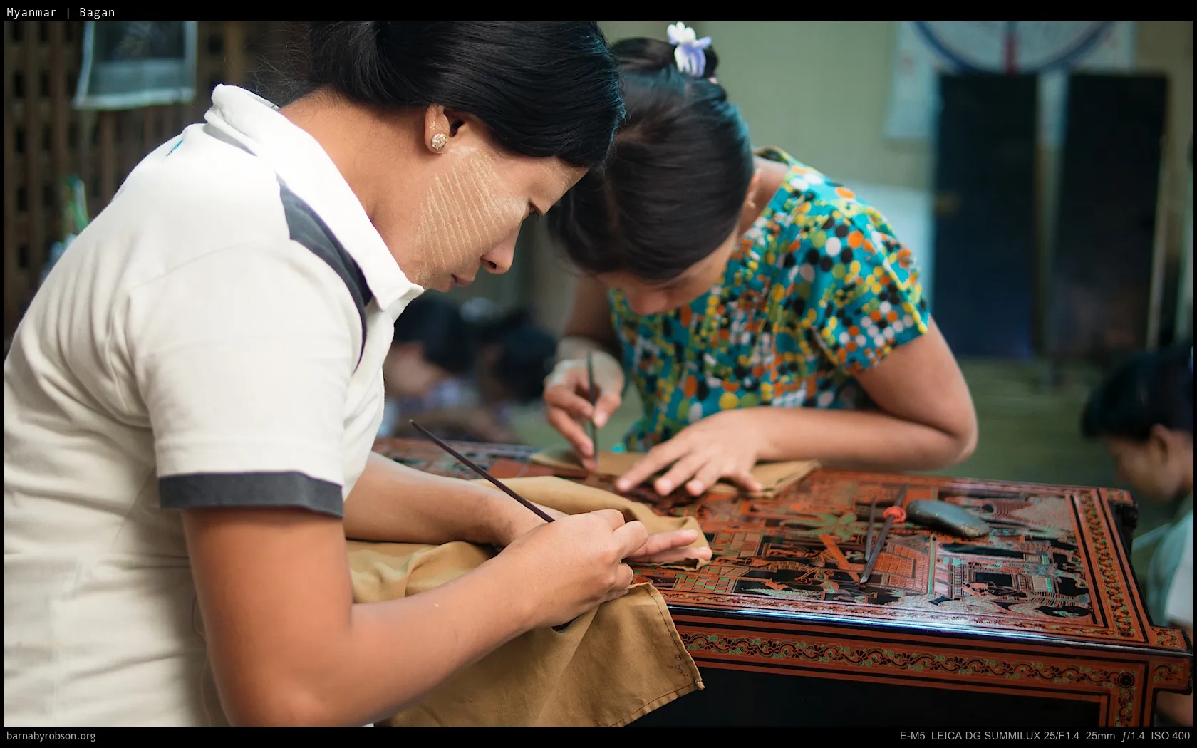 Two women with thanaka face paste crafting at an ornate wooden table.