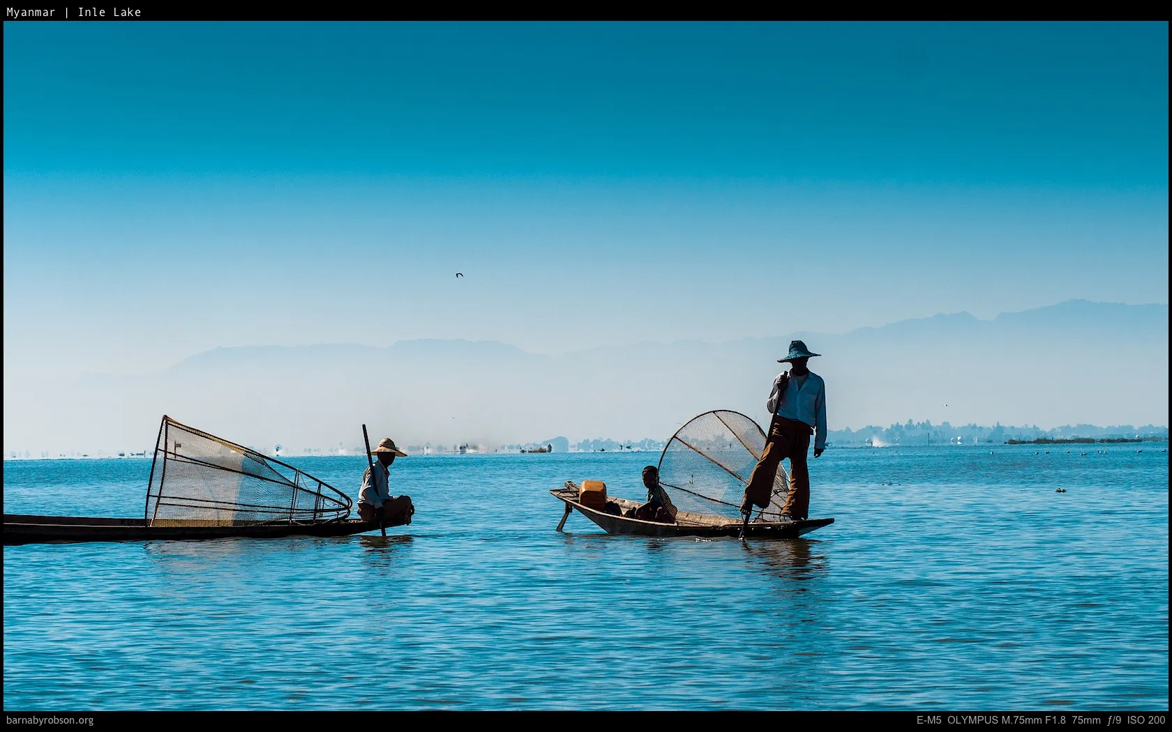 Three people in two boats, one poling with a leg, nets on a lake.