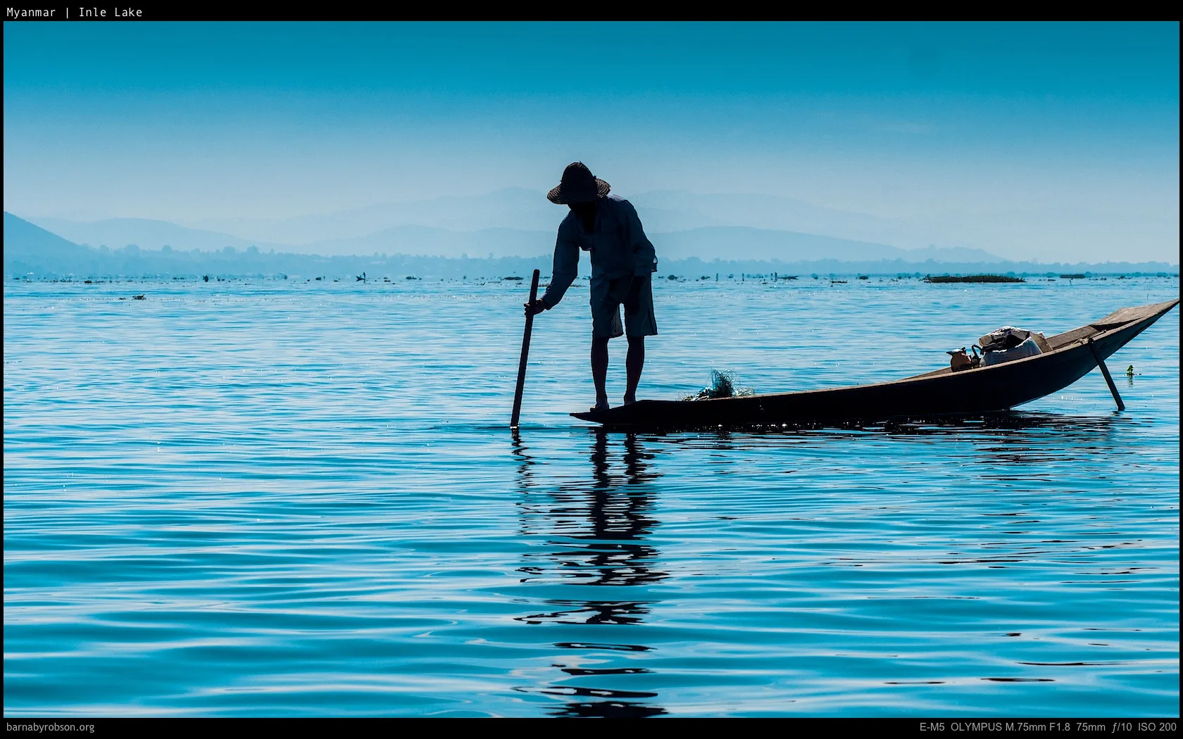 Silhouetted person in a hat poling a wooden boat on a blue lake.