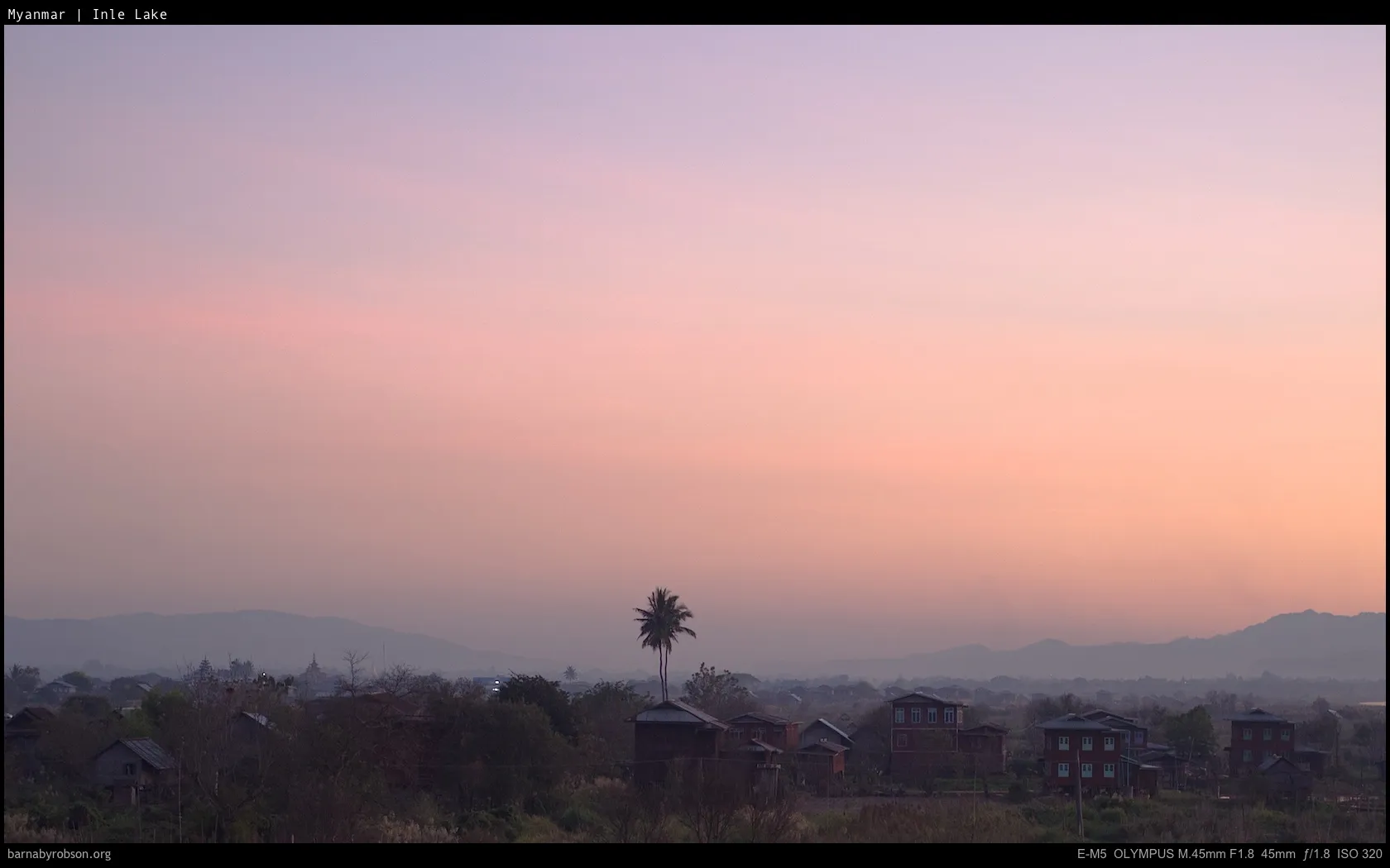 Hazy sunset over a village with houses, trees, and distant mountains.