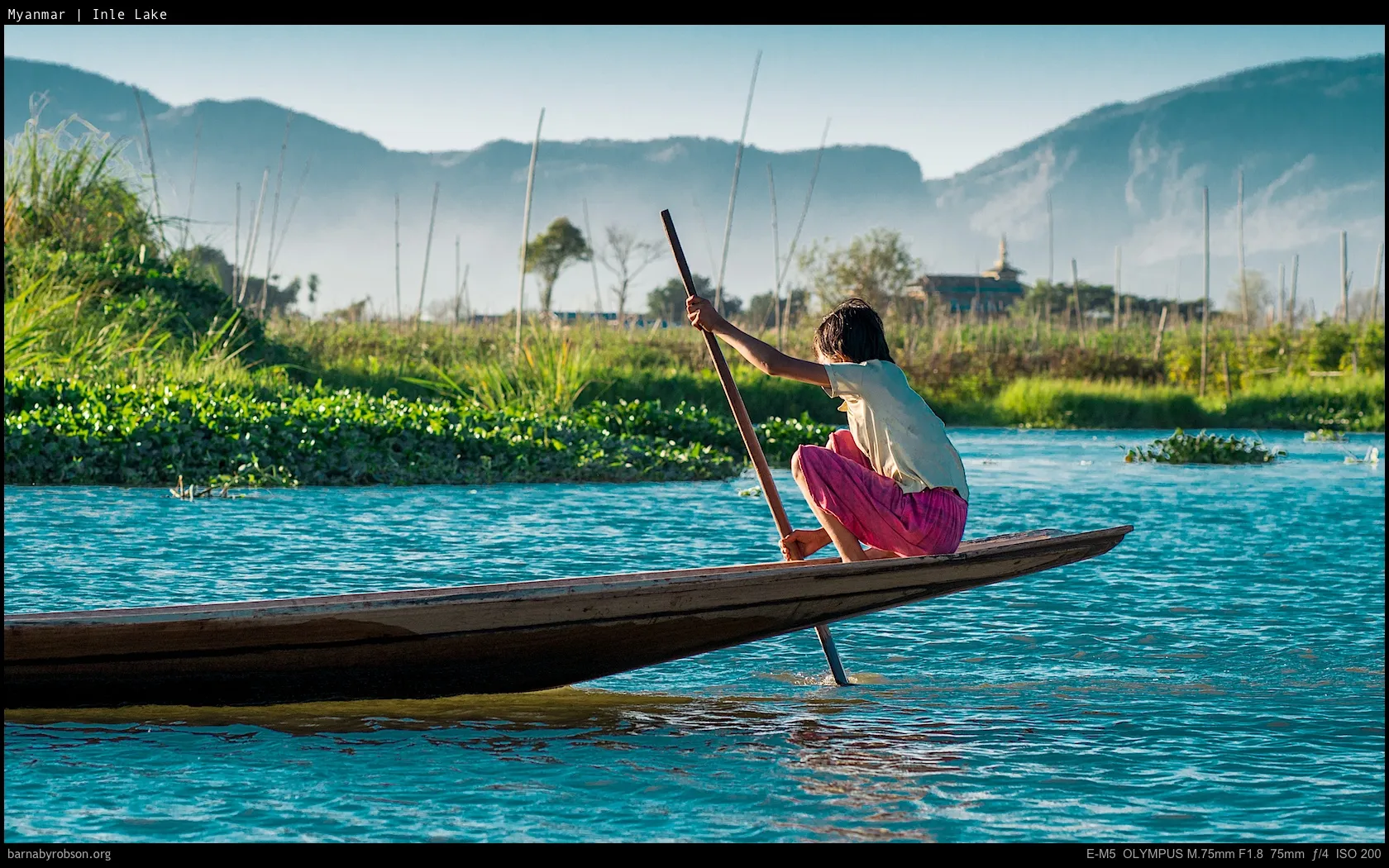 Person poling a wooden boat on blue water, green vegetation, distant mountains.