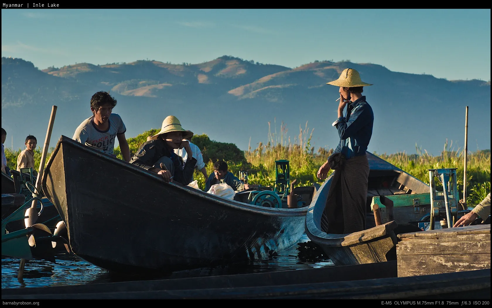 People in wooden boats on water, some wearing straw hats, with distant hills.