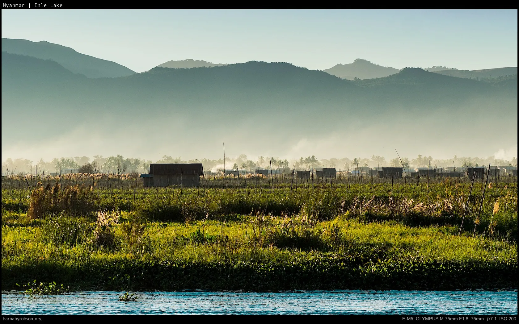 Misty mountains over green fields, wooden huts, and water.