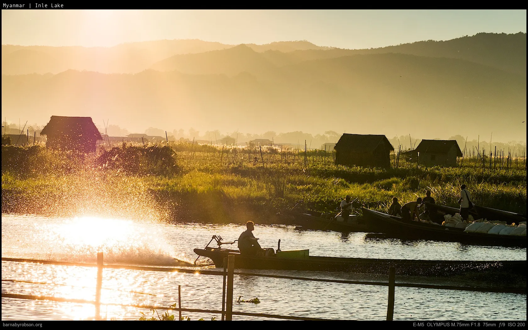 Golden sun illuminates boat splash on lake, with huts and hazy mountains.