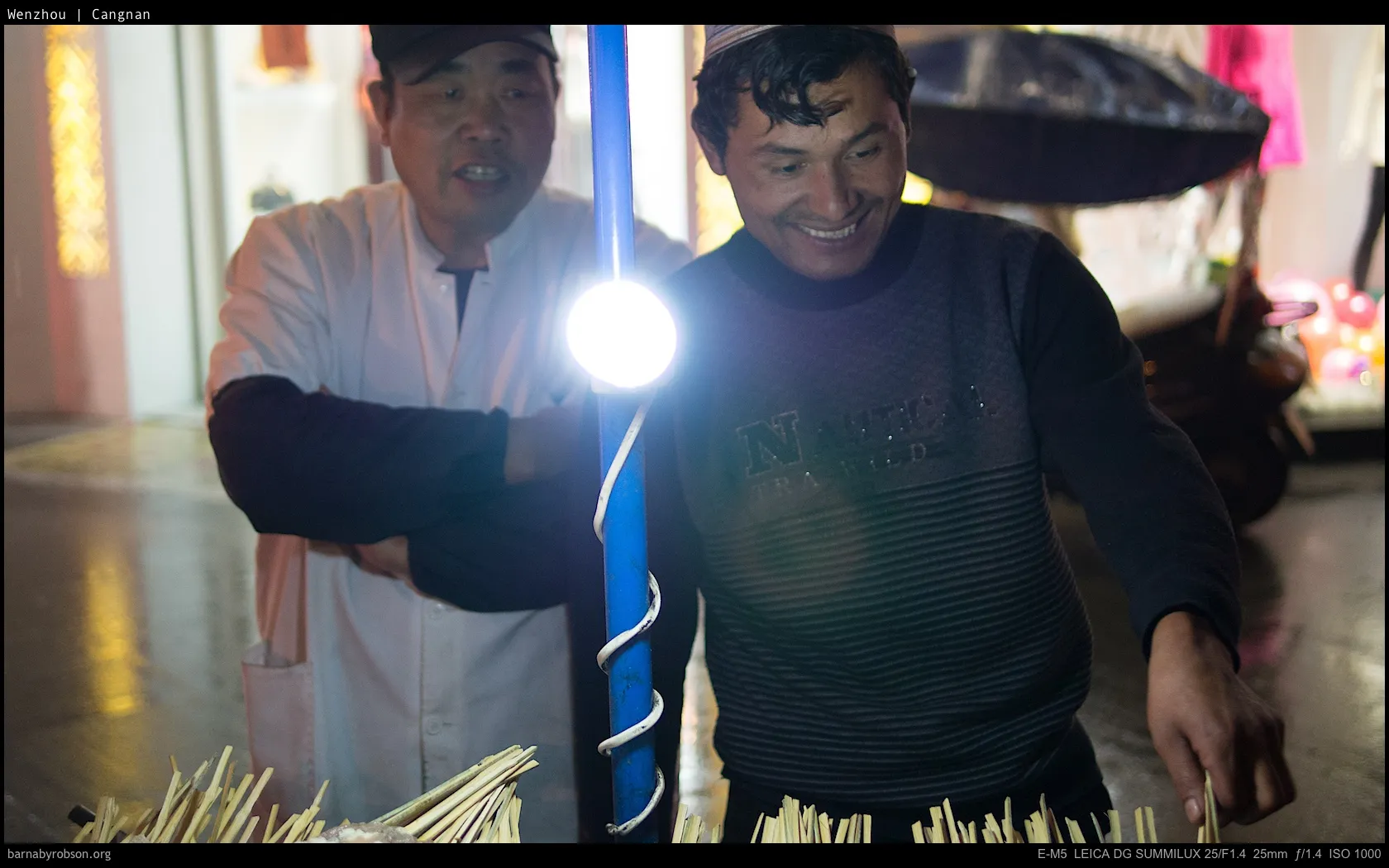 Smiling man with cap, another man, bright light, and wooden skewers.