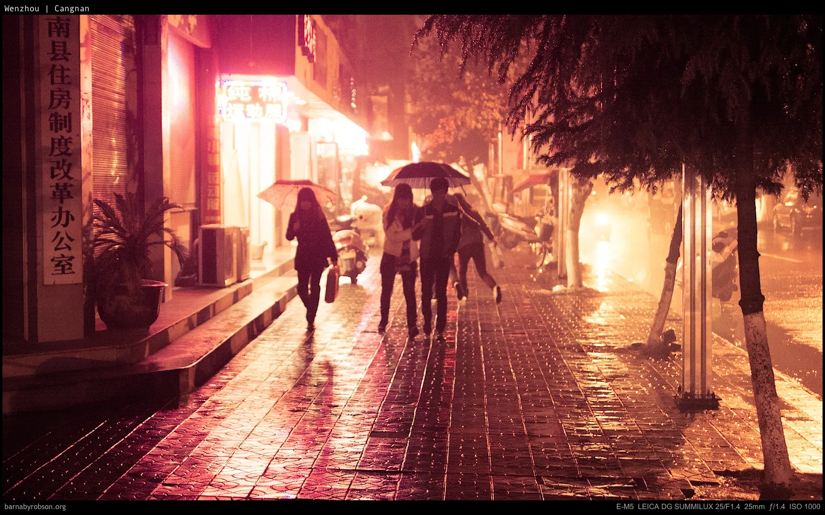 People with umbrellas walk on a wet, glowing street at night.