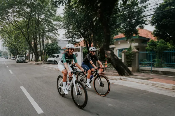 Two cyclists riding side by side down a tree-lined Jakarta street, laughing, tiled roofs and overhead cables behind them.